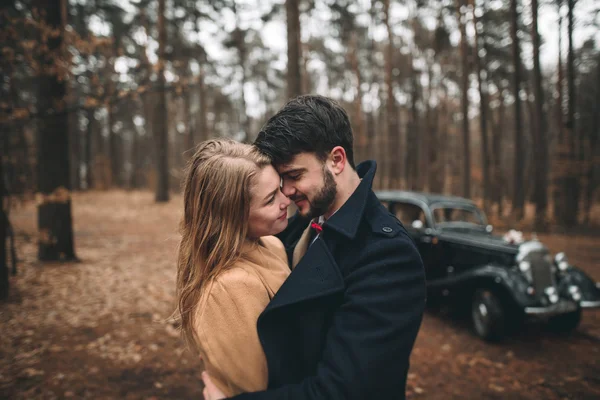 Stylish Loving wedding couple kissing and hugging in a pine forest near retro car — Stock Photo, Image