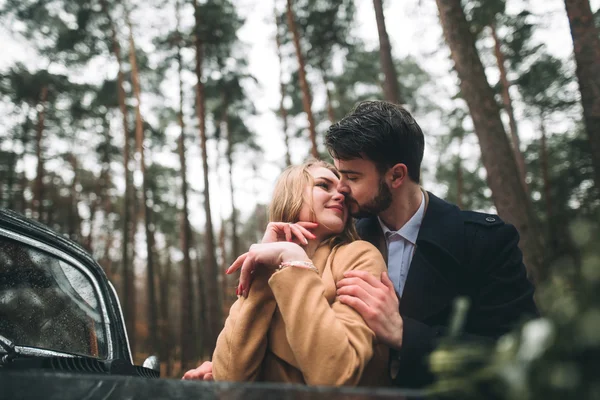 Stylish Loving wedding couple kissing and hugging in a pine forest near retro car — Stock Photo, Image