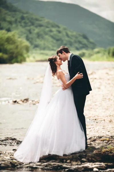 Beautifull wedding couple kissing and embracing near the shore of a mountain river with stones — Stock Photo, Image