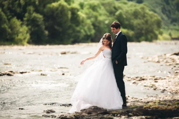 Schönes Hochzeitspaar küsst und umarmt sich am Ufer eines Gebirgsflusses mit Steinen — Stockfoto