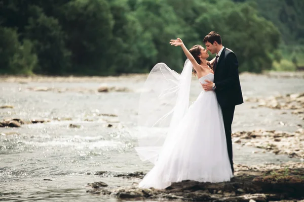 Hermosa pareja de boda besándose y abrazándose cerca de la orilla de un río de montaña con piedras — Foto de Stock
