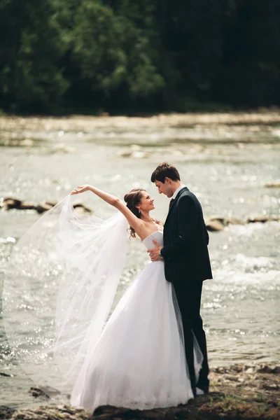 Elegante novio elegante y elegante novia cerca del río con piedras. Pareja de boda enamorada —  Fotos de Stock