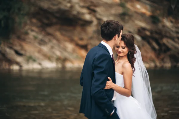 Beautifull wedding couple kissing and embracing near the shore of a mountain river with stones — Stock Photo, Image