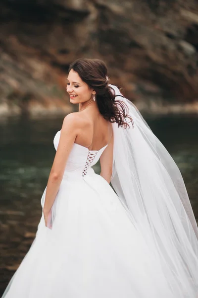 Beautiful luxury young bride in long white wedding dress and veil standing near river with mountains on background — Stock Photo, Image