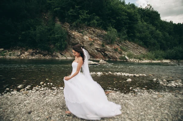 Linda noiva jovem de luxo em vestido de noiva branco longo e véu de pé perto do rio com montanhas no fundo — Fotografia de Stock