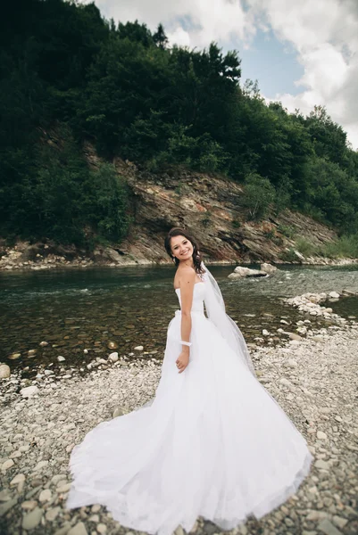 Bella lusso giovane sposa in abito da sposa lungo bianco e velo in piedi vicino al fiume con le montagne sullo sfondo — Foto Stock