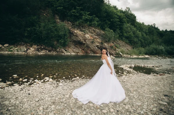 Krásné luxusní mladá nevěsta v dlouhé bílé svatební šaty a závoj stojící poblíž řeky s horami na pozadí — Stock fotografie