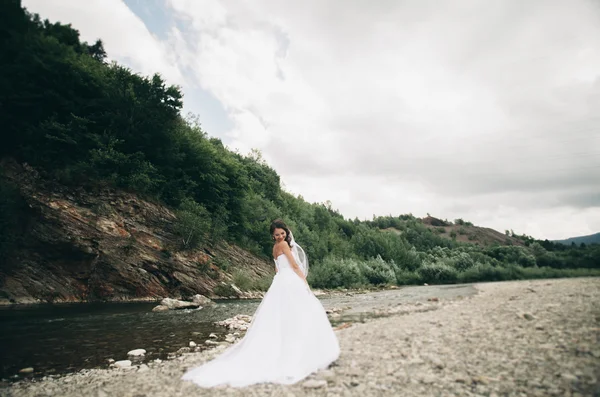 Linda noiva jovem de luxo em vestido de noiva branco longo e véu de pé perto do rio com montanhas no fundo — Fotografia de Stock
