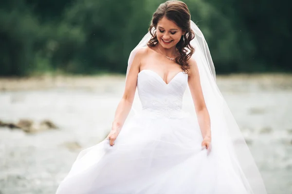 Hermosa novia joven de lujo en vestido de novia largo y blanco y velo de pie cerca del río con montañas en el fondo — Foto de Stock