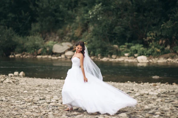 Krásné luxusní mladá nevěsta v dlouhé bílé svatební šaty a závoj stojící poblíž řeky s horami na pozadí — Stock fotografie