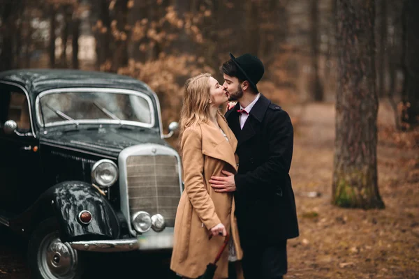 Romantisches Märchenhochzeitspaar küsst und umarmt sich im Kiefernwald in der Nähe des Retro-Autos. — Stockfoto