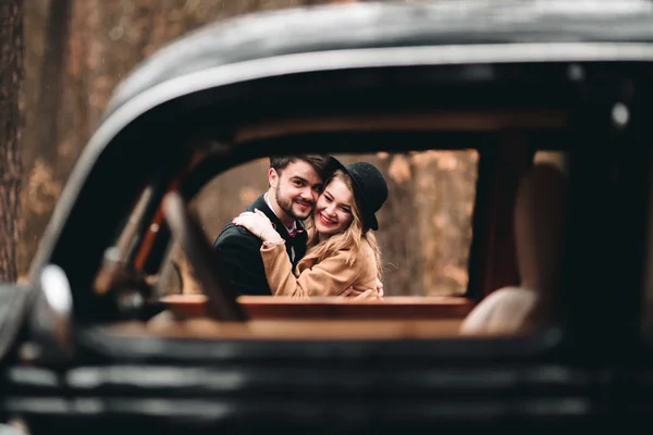 Romántica pareja de boda de cuento de hadas besándose y abrazándose en el bosque de pinos cerca de coche retro . — Foto de Stock