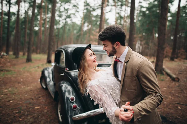 Романтическая сказочная свадебная пара целуется и обнимается в сосновом лесу рядом с ретро-машиной . — стоковое фото