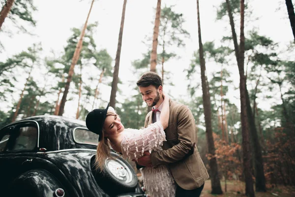 Romantic fairytale wedding couple kissing and embracing in pine forest near retro car. — Stock Photo, Image