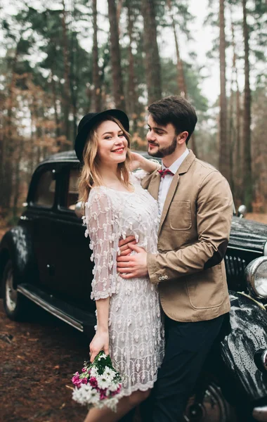 Romantic fairytale wedding couple kissing and embracing in pine forest near retro car. — Stock Photo, Image