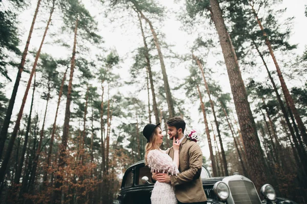 Romantisches Märchenhochzeitspaar küsst und umarmt sich im Kiefernwald in der Nähe des Retro-Autos. — Stockfoto