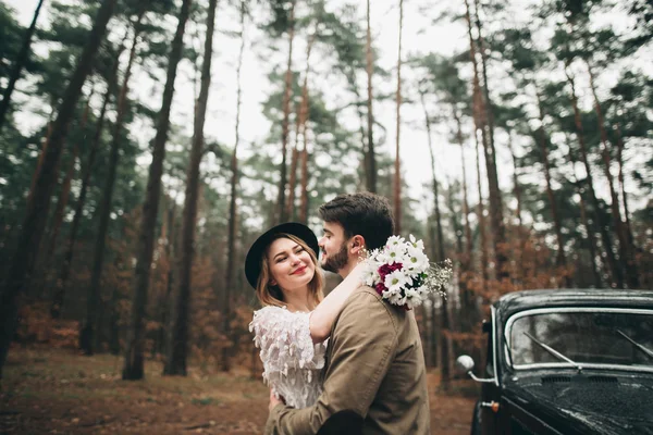 Romantisches Märchenhochzeitspaar küsst und umarmt sich im Kiefernwald in der Nähe des Retro-Autos. — Stockfoto