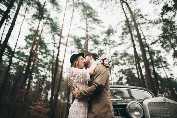 Romantic fairytale wedding couple kissing and embracing in pine forest near retro car. — Stock Photo, Image