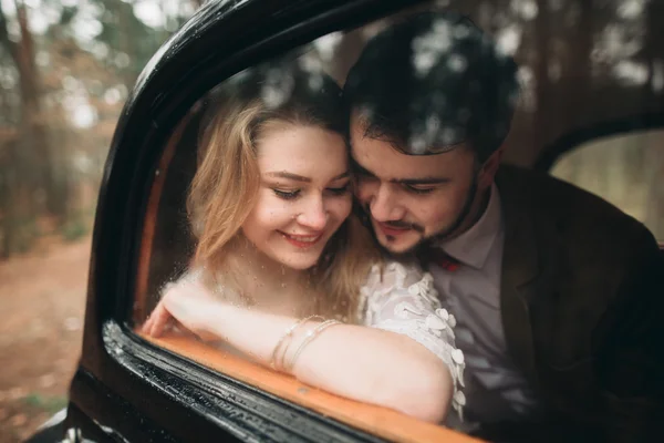 Romantikus mese esküvői pár csók, és átfogó, a fenyves erdő közelében retro autó. — Stock Fotó