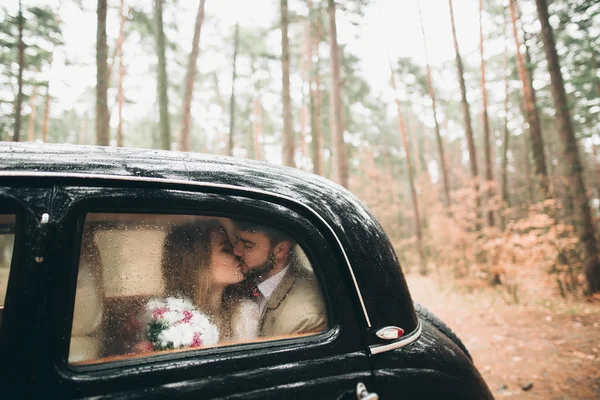 Romantic fairytale wedding couple kissing and embracing in pine forest near retro car. — Stock Photo, Image