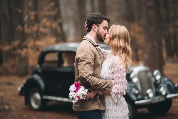 Romantic fairytale wedding couple kissing and embracing in pine forest near retro car. — Stock Photo, Image