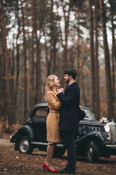 Romantic fairytale wedding couple kissing and embracing in pine forest near retro car. — Stock Photo, Image