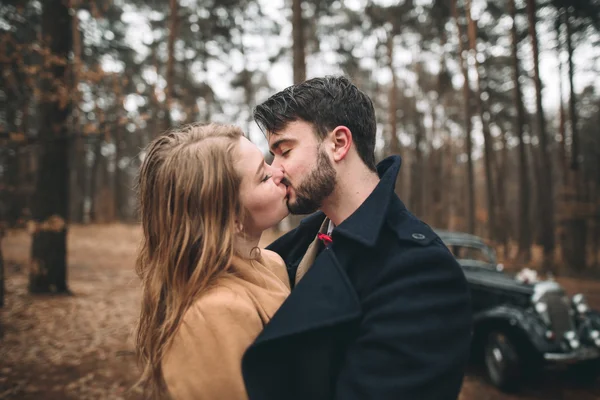 Romantikus mese esküvői pár csók, és átfogó, a fenyves erdő közelében retro autó. — Stock Fotó