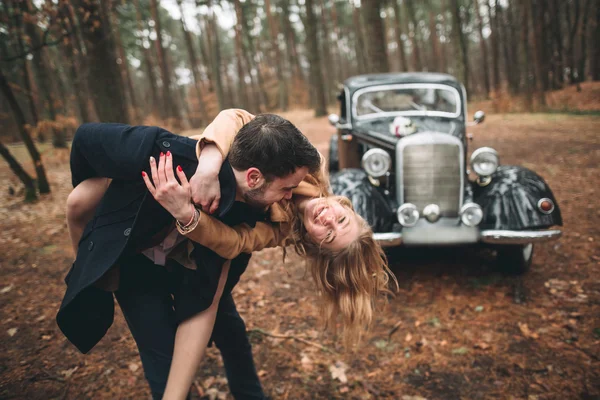 Romántica pareja de boda de cuento de hadas besándose y abrazándose en el bosque de pinos cerca de coche retro . — Foto de Stock