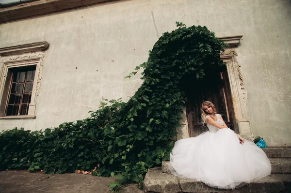 Belle jeune mariée blonde avec bouquet nuptial assis sur les escaliers sur le fond de magnifiques plantes — Photo