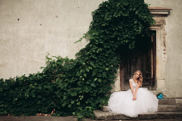 Krásná mladá blonďatá nevěsta s svatební kytice sedí na schodech na pozadí nádherné rostliny — Stock fotografie