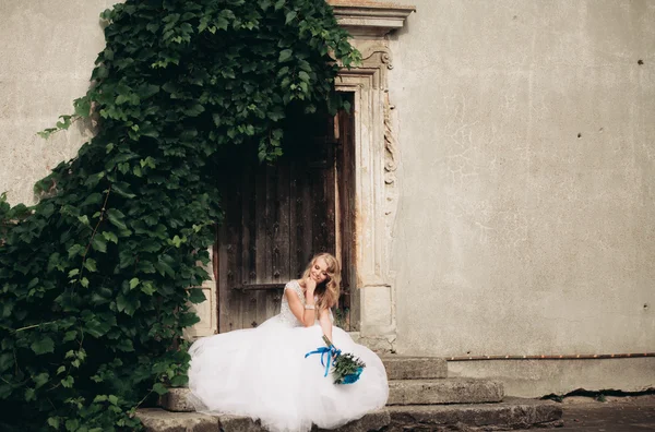 Belle jeune mariée blonde avec bouquet nuptial assis sur les escaliers sur le fond de magnifiques plantes — Photo