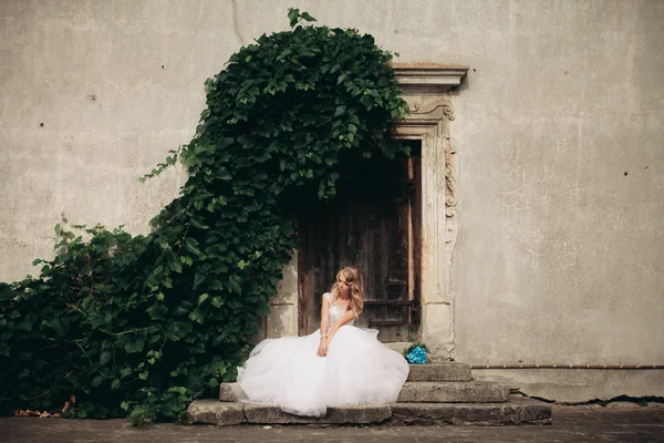 Belle jeune mariée blonde avec bouquet nuptial assis sur les escaliers sur le fond de magnifiques plantes — Photo