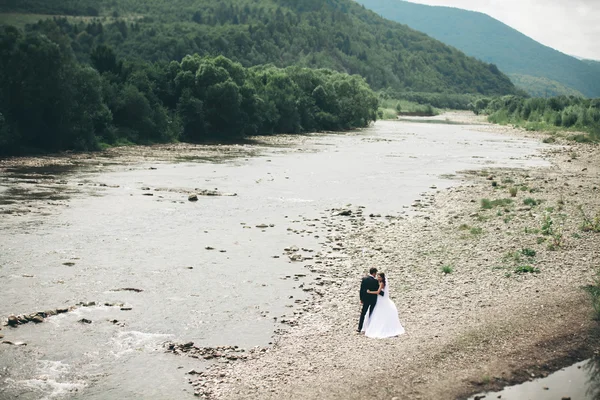 Bellissimo matrimonio coppia baciare e abbracciare vicino alla riva di un fiume di montagna con pietre — Foto Stock