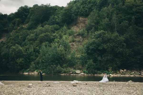 Hermosa pareja de boda besándose y abrazándose cerca de la orilla de un río de montaña con piedras — Foto de Stock