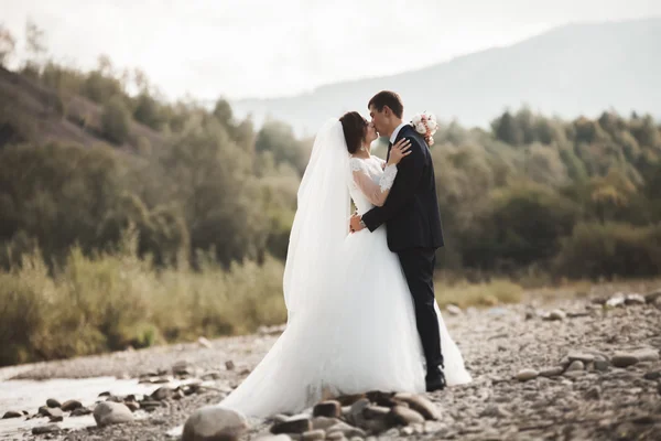 Guapo romántico novio y hermosa novia posando cerca del río en las montañas escénicas — Foto de Stock