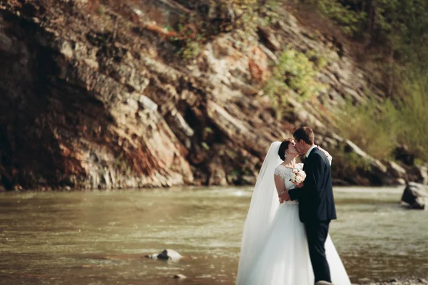 Handsome romantic groom and beautiful bride posing near river in scenic mountains — Stock Photo, Image