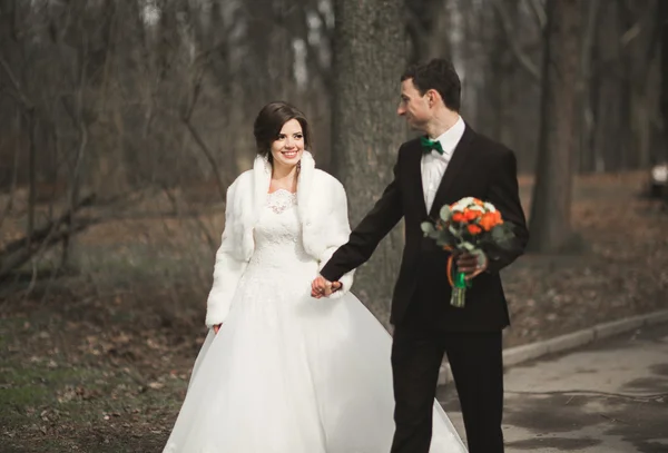 Красивая пара счастливых стильных молодоженов, прогуливающихся в парке в день своей свадьбы с букетом — стоковое фото