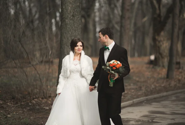 Красивая пара счастливых стильных молодоженов, прогуливающихся в парке в день своей свадьбы с букетом — стоковое фото