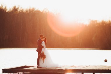 Zarif güzel düğün çifti bir göl kenarında günbatımında poz