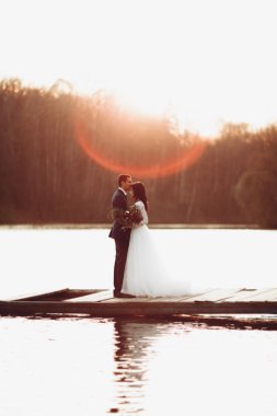 Zarif güzel düğün çifti bir göl kenarında günbatımında poz