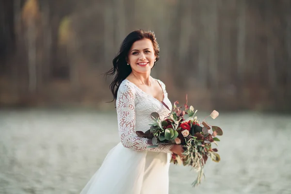Linda noiva morena em vestido elegante segurando buquê posando perto da floresta e do lago — Fotografia de Stock