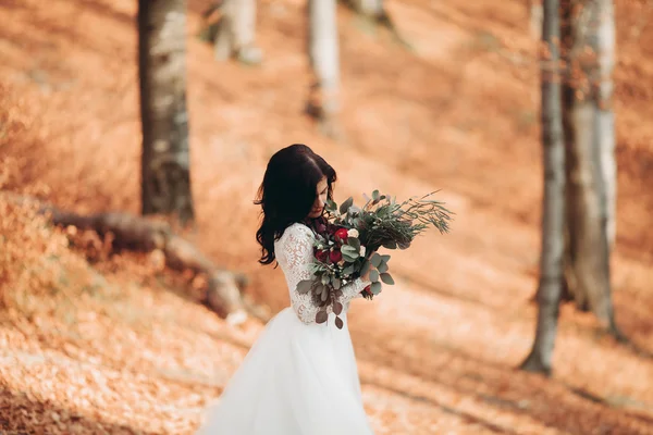 Splendida sposa bruna in elegante vestito che tiene bouquet in posa vicino alla foresta e al lago — Foto Stock