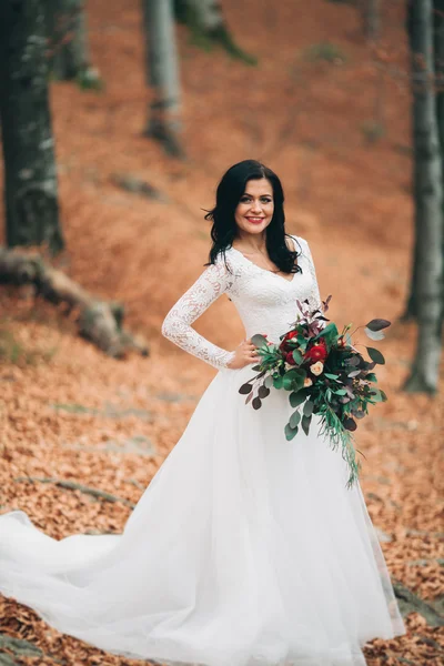 Hermosa novia morena en vestido elegante celebración de ramo posando cerca del bosque y el lago — Foto de Stock