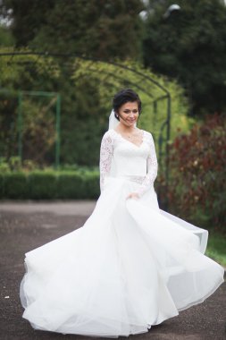 Zarif beyaz elbiseli güzel esmer gelin elinde buket ile güzel ağaçlar poz veriyor.