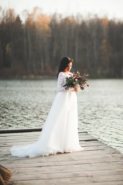 Prachtige brunette bruid in elegante jurk bedrijf boeket poseren in de buurt van bos en meer — Stockfoto