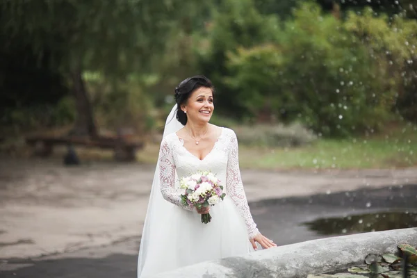 Beautiful brunette bride in elegant white dress holding bouquet posing neat trees — Stock Photo, Image