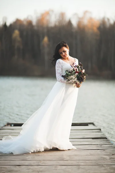 Prachtige brunette bruid in elegante jurk bedrijf boeket poseren in de buurt van bos en meer — Stockfoto