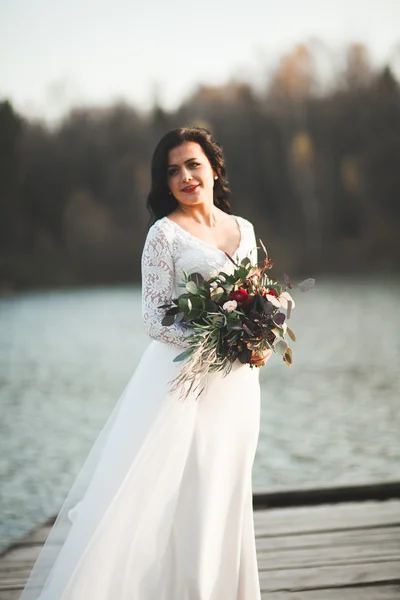 Vacker brunett brud i elegant klänning holding bukett poserar nära skog och sjö — Stockfoto