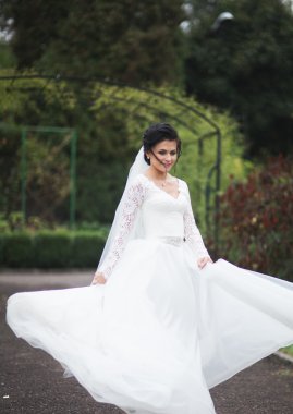 Zarif beyaz elbiseli güzel esmer gelin elinde buket ile güzel ağaçlar poz veriyor.