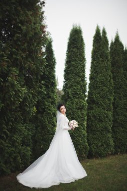 Zarif beyaz elbiseli güzel esmer gelin elinde buket ile güzel ağaçlar poz veriyor.